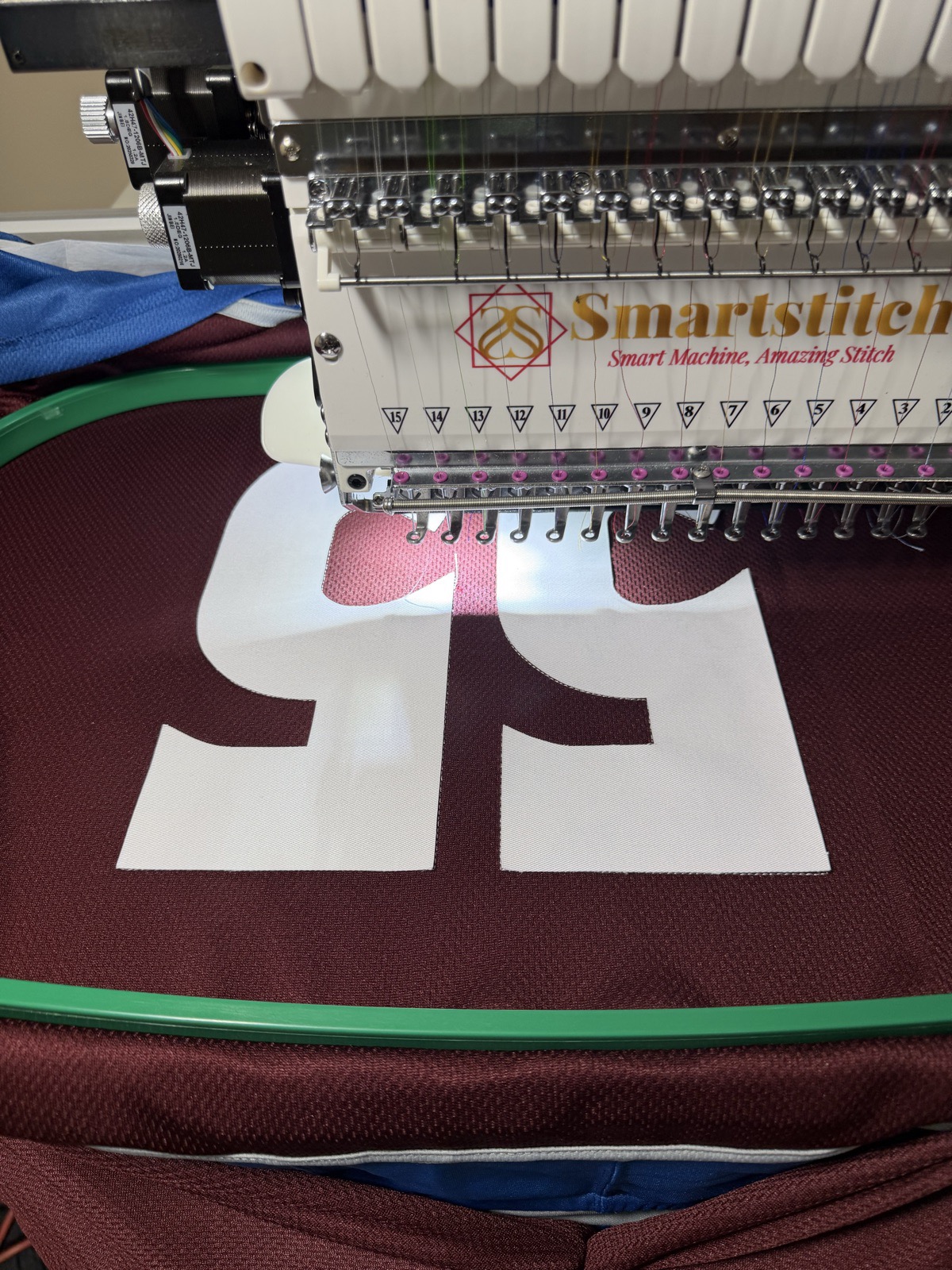 Smartstitch multi-needle embroidery machine in action, stitching white tackle-twill SPUTNIK lettering onto maroon hoodie fabric