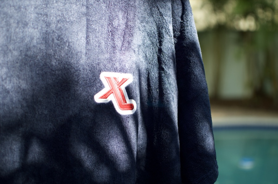 Xeno Pucks branded hooded poncho with embroidered X patch close-up and pool bokeh background