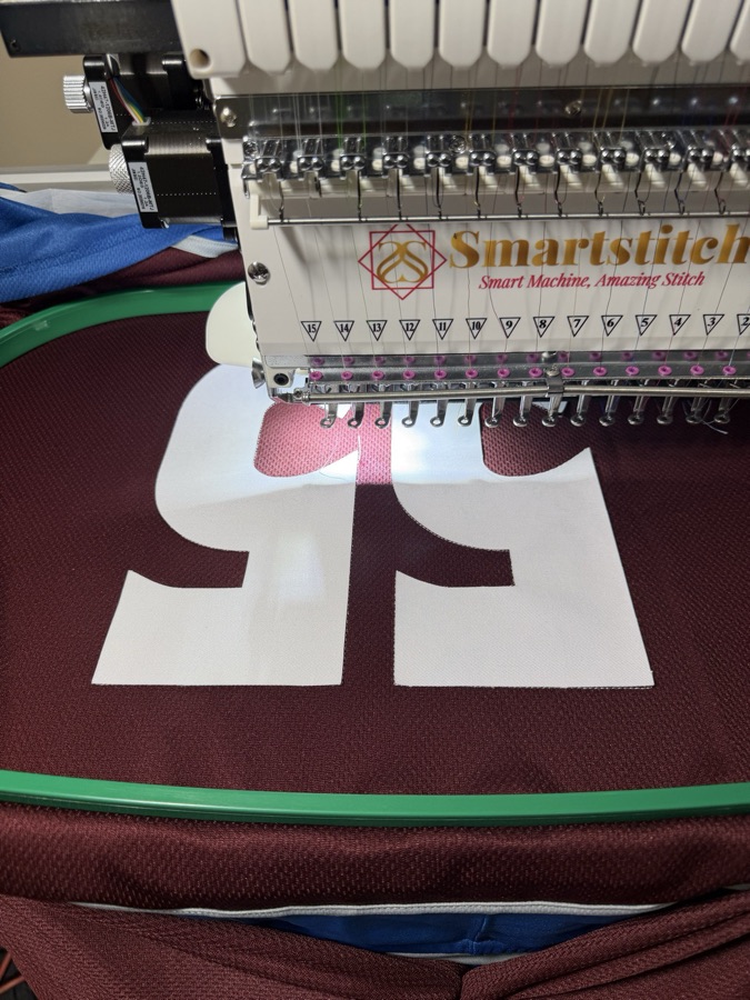 Smartstitch multi-needle embroidery machine in action, stitching white tackle-twill SPUTNIK lettering onto maroon hoodie fabric